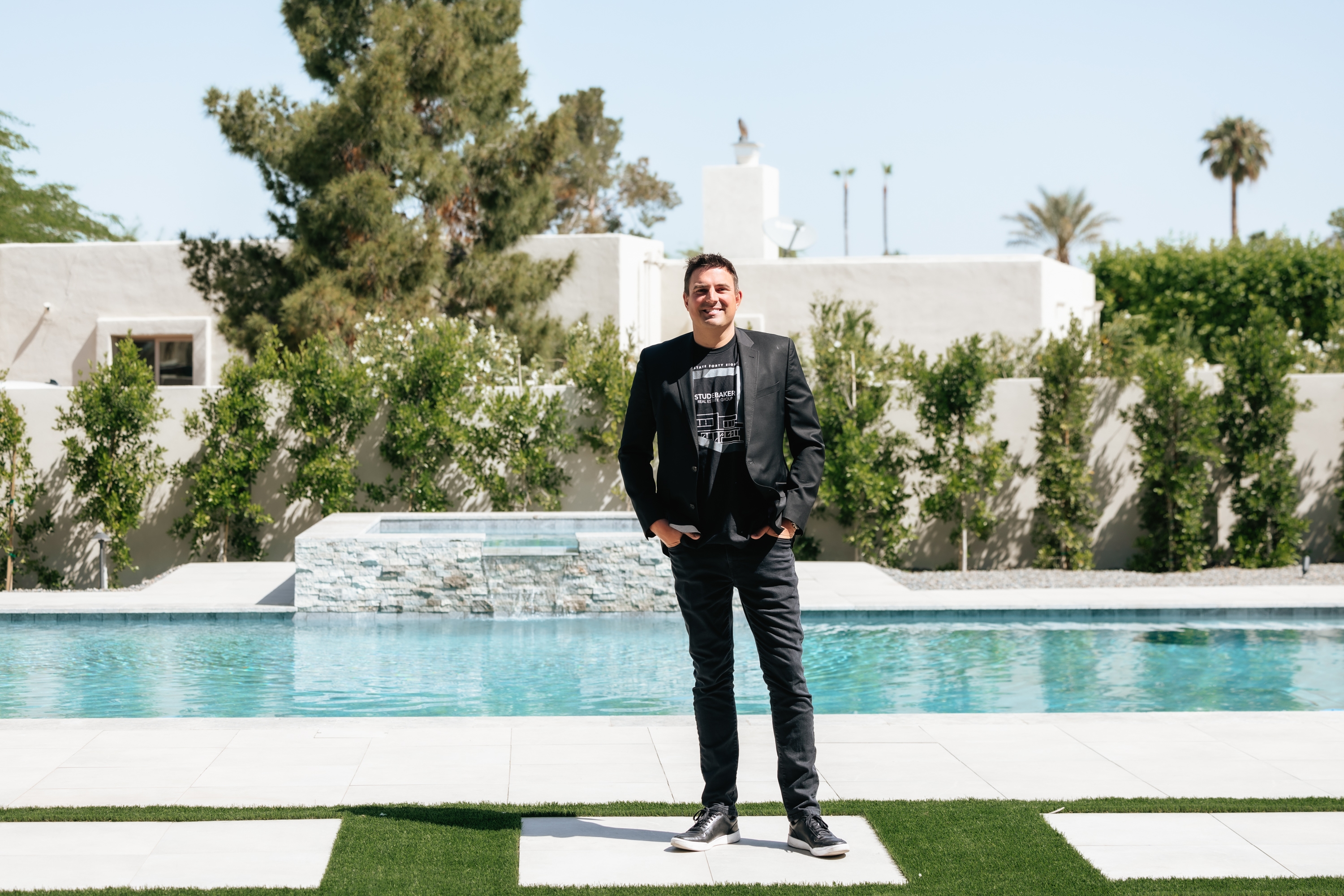 A smiling man in a black blazer and a "Studebaker Real Estate Group" t-shirt stands by a modern swimming pool with a tiled patio, surrounded by white walls and greenery.