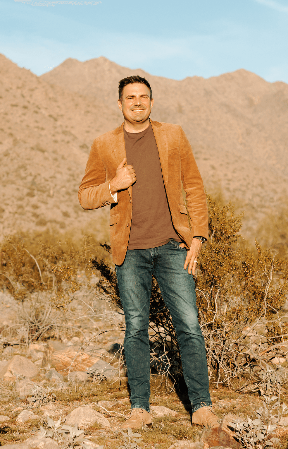 A smiling man in a black suit and sneakers leans casually against a white doorway in a bright, modern interior.