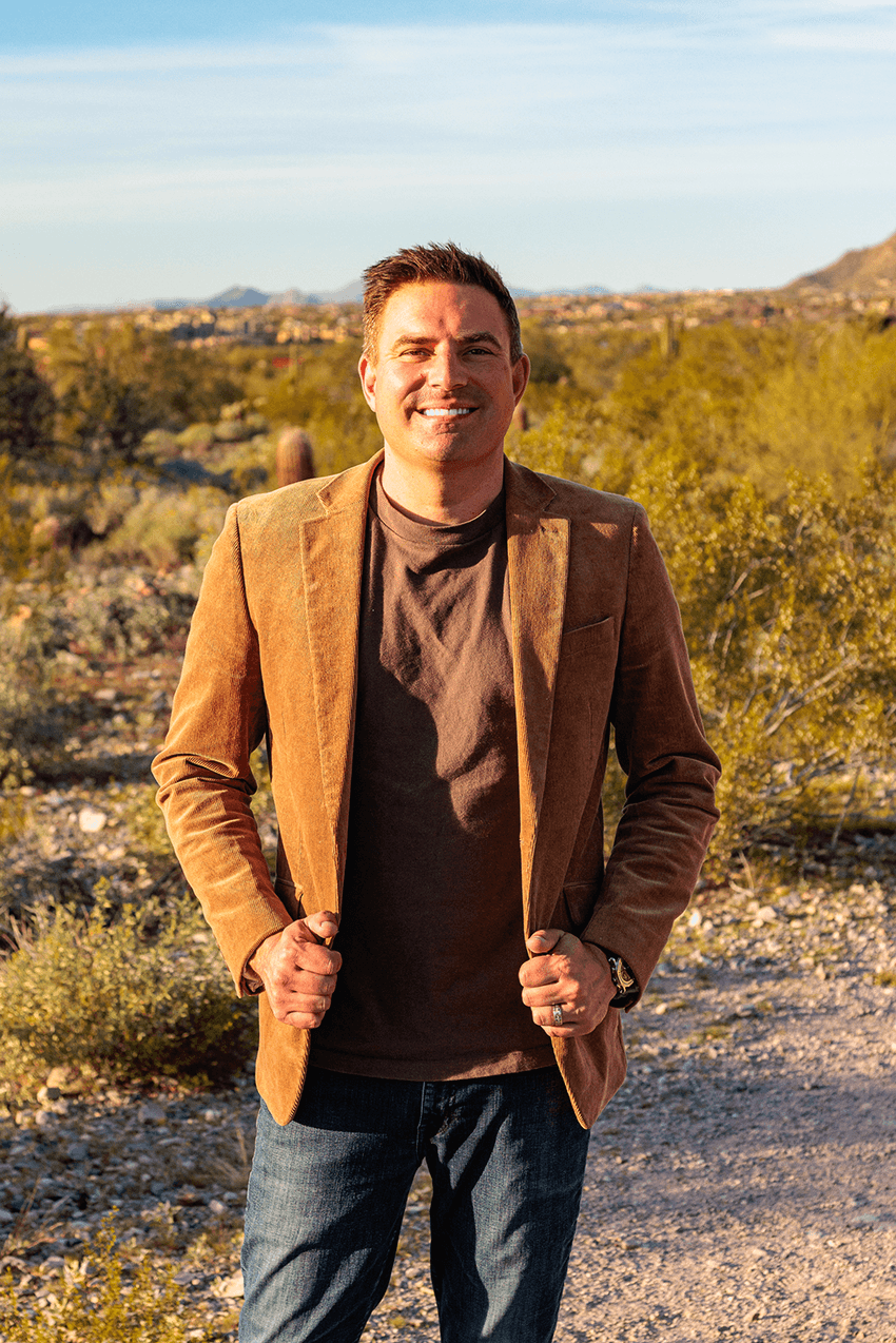 A smiling man in a black suit and sneakers leans casually against a white doorway in a bright, modern interior.