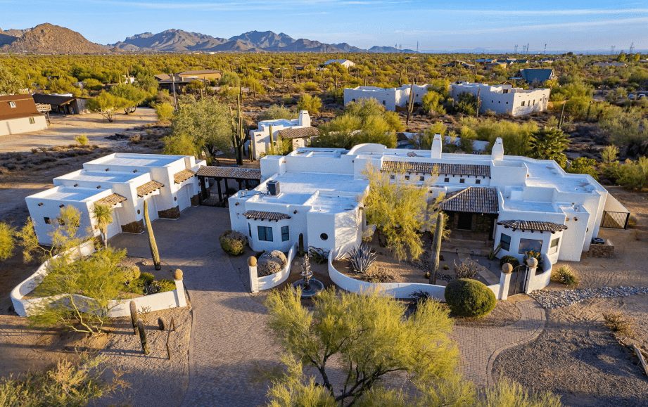 Aerial view of a large white Southwestern-style house with red tile roofs, set in a desert landscape with saguaro cacti and distant mountains under a blue sky.