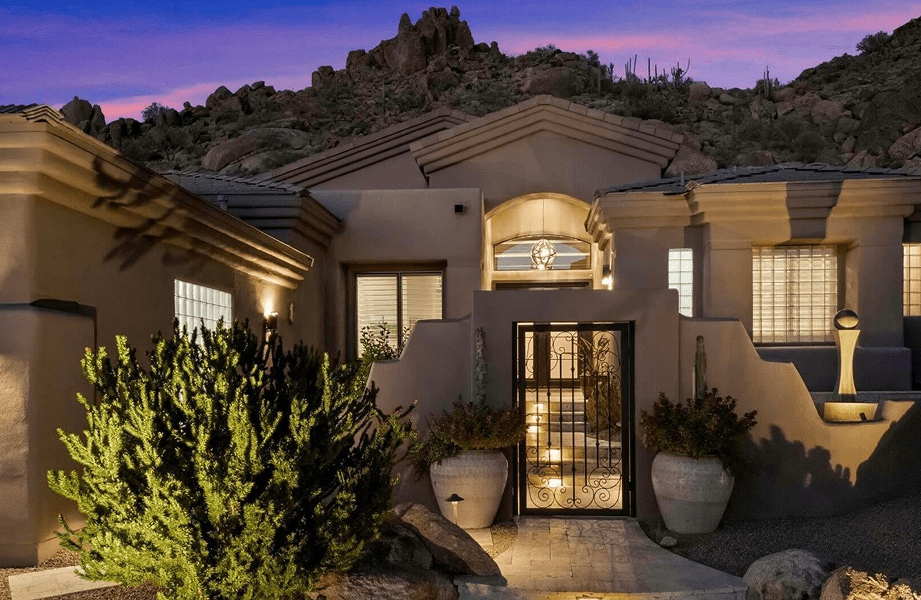 Elegant desert home exterior at twilight, illuminated by warm lights, featuring an ornate iron gate, landscaped entryway, and rocky mountains under a vibrant purple sky.