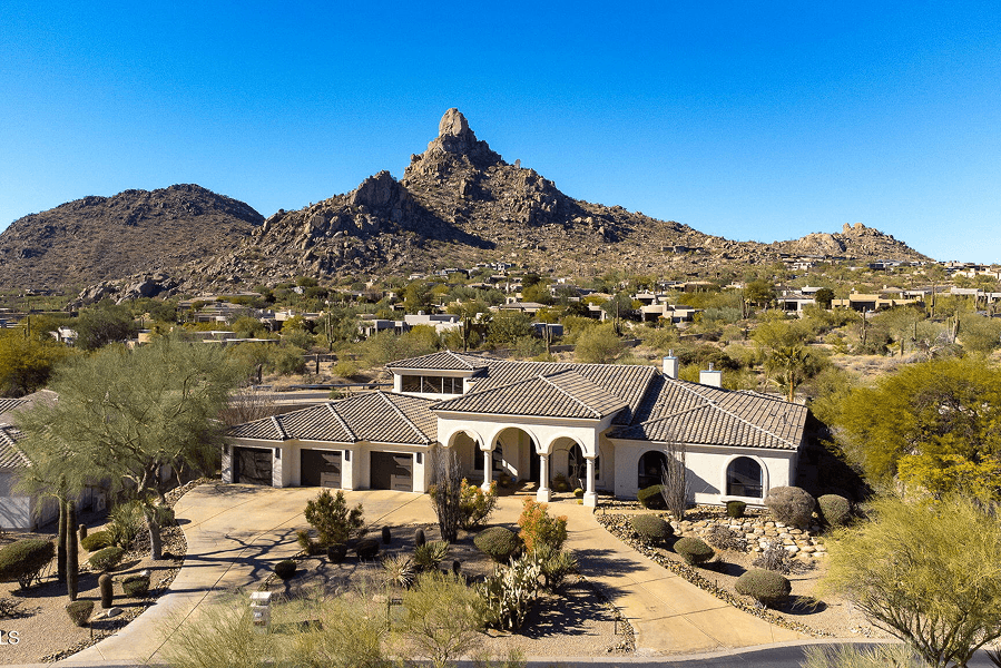An aerial view of a large, cream-colored house with a tiled roof and circular driveway, set amidst desert landscaping with rugged mountains and a prominent rocky peak under a clear blue sky.