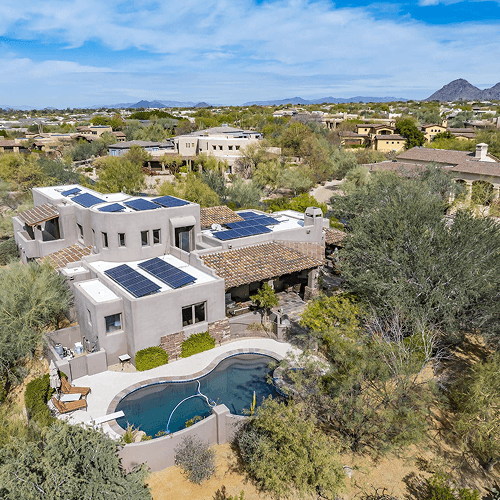 Aerial view of a modern desert home with numerous solar panels, a kidney-shaped pool, and arid landscaping, surrounded by other houses and distant mountains.