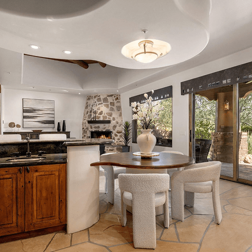 An elegant open-plan living area featuring a kitchen island, a round wooden dining table with modern white chairs, a stone fireplace, and a bright view to an outdoor patio.