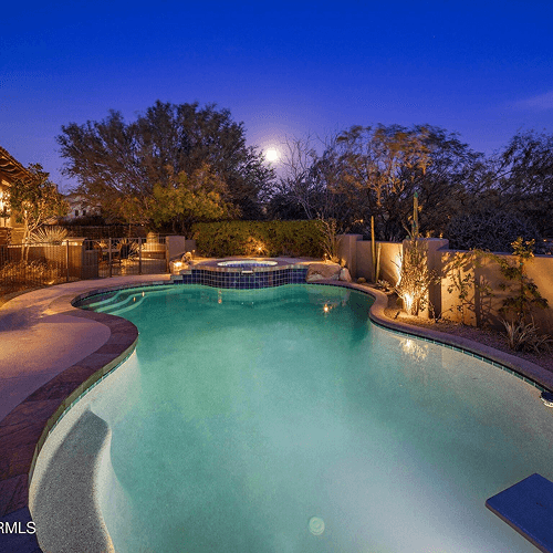 An illuminated swimming pool and hot tub in a landscaped desert backyard under a clear blue night sky with a full moon.