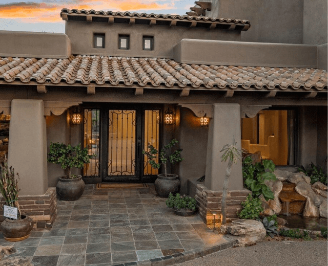 Southwestern-style home entrance at sunset with terracotta tiled roof, stucco walls, ornate double doors, potted plants, and a small waterfall feature.