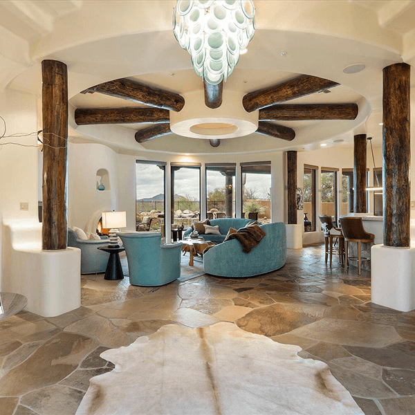 A grand circular living space with rustic log columns supporting the ceiling, teal furniture, a cowhide rug on flagstone flooring, and large windows revealing a desert view.