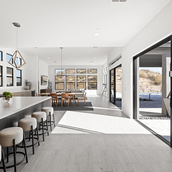 A bright, modern open-concept living space featuring a kitchen island with stools, dining area, and living room, all illuminated by large windows overlooking a desert landscape. A large sliding glass door opens to an outdoor patio.