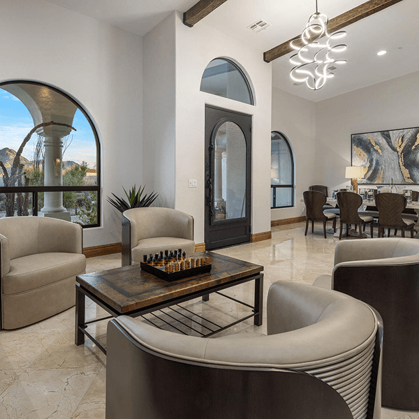 Luxurious living room featuring a chess set on a coffee table, surrounded by light armchairs, with arched windows overlooking mountains, and a modern chandelier illuminating a distant dining area.