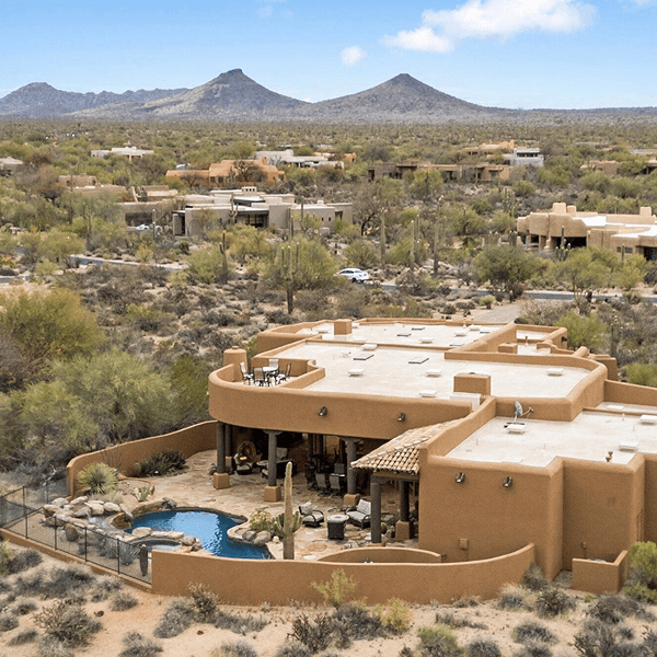 Aerial view of an adobe-style house with a pool and patio, nestled in a desert landscape with saguaro cacti and distant mountains under a blue sky.