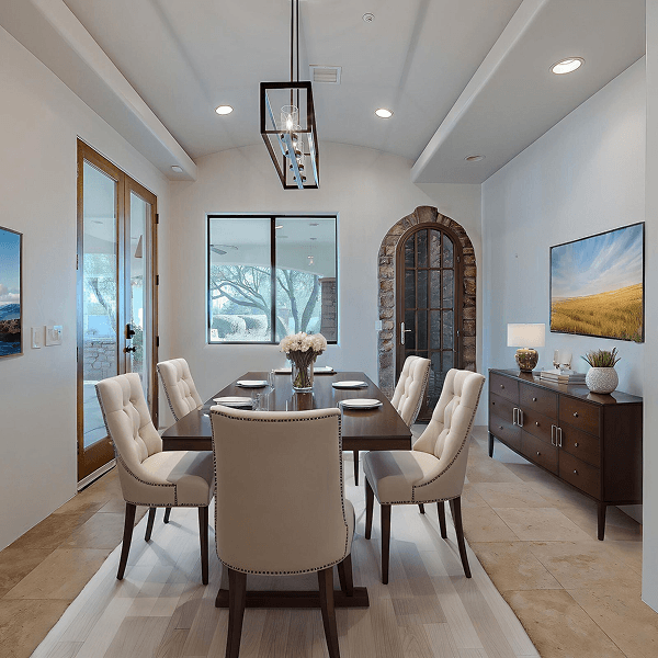 An elegant dining room featuring a long dark wooden table surrounded by cream upholstered chairs, a modern chandelier, and a buffet with a flat-screen TV on the wall. Large windows and a stone arched doorway add character.