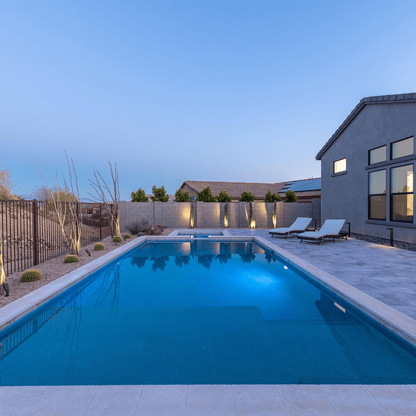 A modern backyard features a glowing blue swimming pool, a spa, and a light-colored patio next to a house. Desert landscaping with illuminated plants and a block wall complete the serene evening scene.