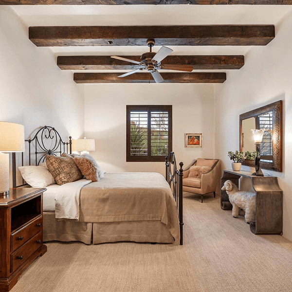 A rustic-style bedroom featuring a wrought iron bed, exposed dark wood ceiling beams, a ceiling fan, a window with plantation shutters, and a cozy sitting area with an armchair.