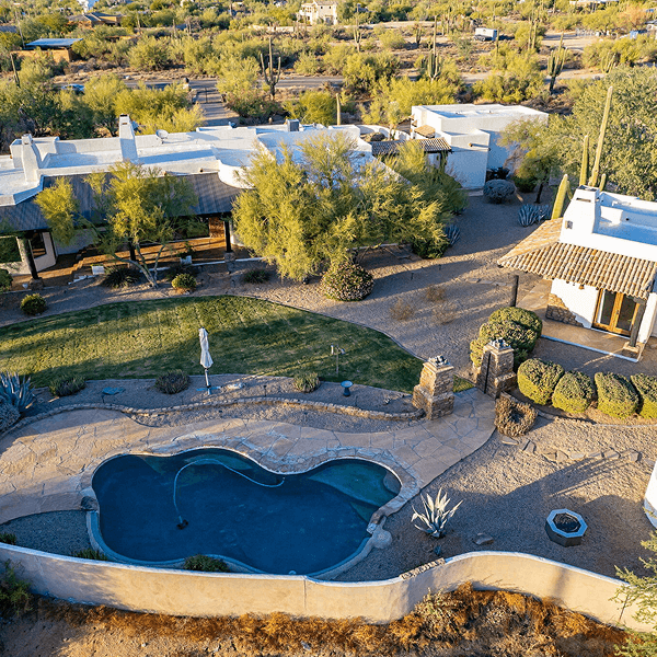 Aerial view of a sprawling desert home featuring white buildings, a freeform swimming pool, a manicured lawn, and arid landscaping with saguaro cacti.
