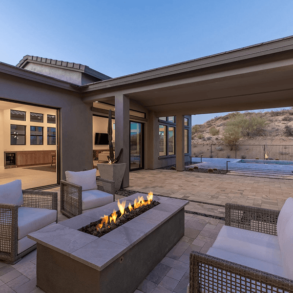 Modern luxury home patio with a gas fire pit, comfortable outdoor seating, open sliding doors to an interior, and a swimming pool overlooking a desert landscape.