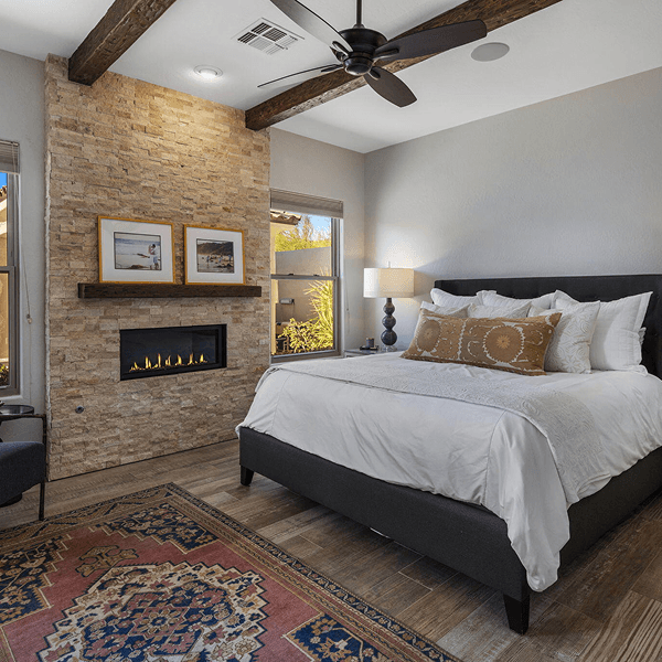 A modern, light-filled bedroom featuring a king-size bed, a stone fireplace accent wall with a wooden mantel, exposed ceiling beams, and a colorful patterned rug on wood-look tile floors.
