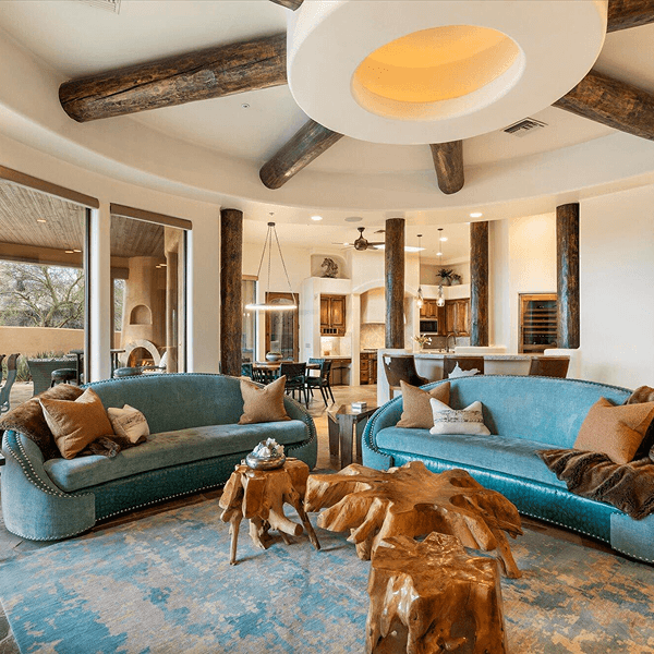A luxurious, open-concept living room with curved teal sofas, organic wood coffee tables, a patterned rug, rustic ceiling beams radiating from a large circular light, and an adjacent kitchen and outdoor patio.
