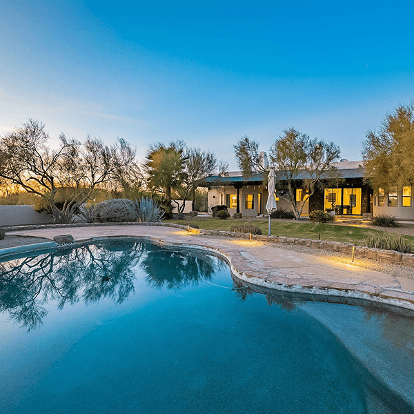 A luxury desert home with a large swimming pool and illuminated patio at dusk, surrounded by sparse trees and desert plants.