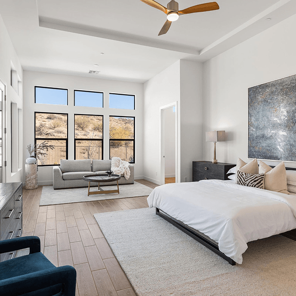A modern, spacious bedroom featuring a white bed, an abstract painting, a sitting area with a grey sofa, and large windows overlooking a desert landscape.