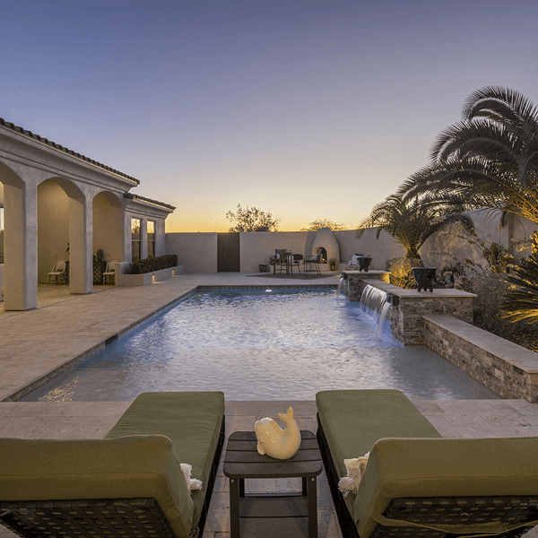 A luxurious backyard features a swimming pool with lit waterfalls, lounge chairs, a house with a patio, and an outdoor fireplace under a twilight sky.