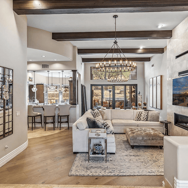 Luxurious open-concept living room and kitchen featuring high ceilings with wood beams, a large sectional sofa, ottoman, grand chandelier, fireplace, and a kitchen island with bar seating.