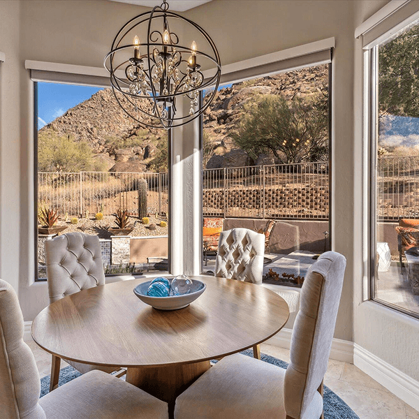 A dining room with a round wooden table and four upholstered chairs, under a spherical chandelier. Large windows offer a sunlit view of a desert mountain landscape and an outdoor patio.