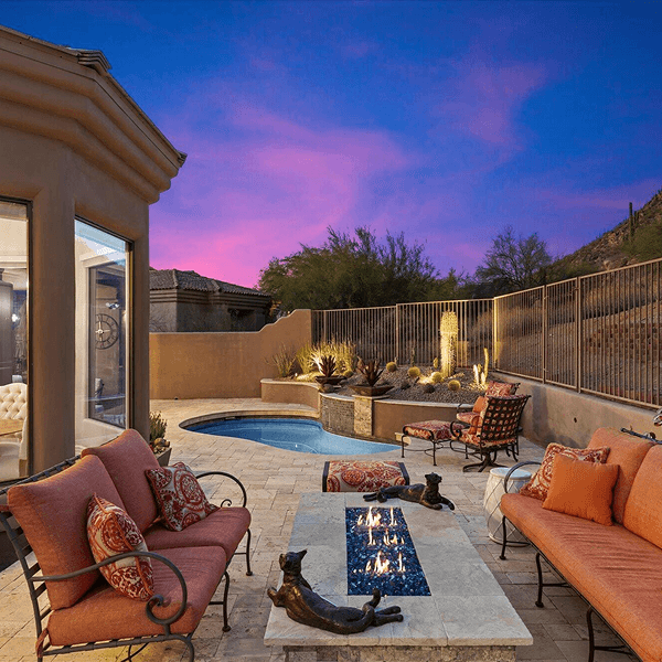 Luxurious desert patio at dusk featuring a glowing fire pit, comfortable orange sofas, a small spa pool, and desert landscaping under a vibrant purple sunset.