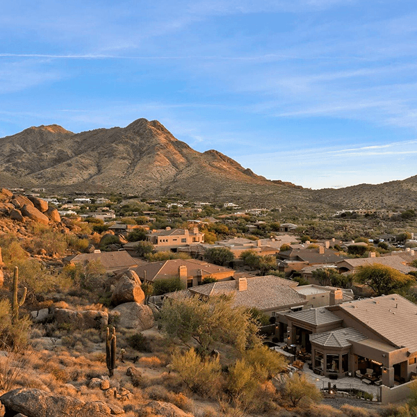 Warm sunlight illuminates a desert landscape with large mountains overlooking a community of modern homes, saguaro cacti, and arid shrubs under a blue sky.
