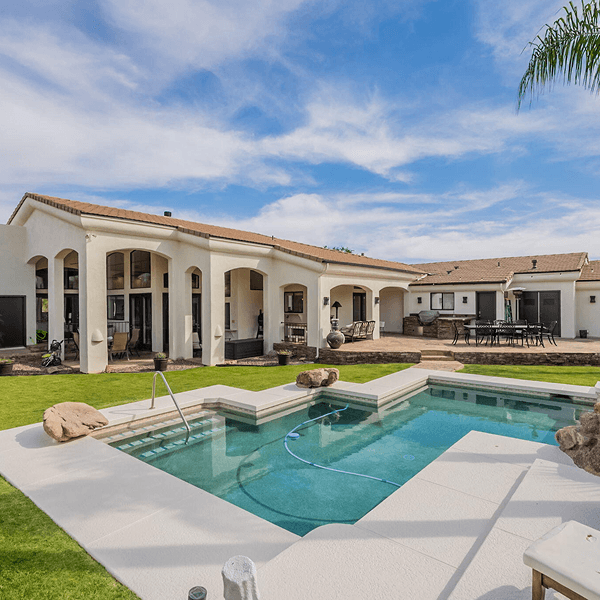 A spacious backyard featuring a large, light-colored house with multiple covered patios, a swimming pool, and an outdoor dining area on a sunny day.