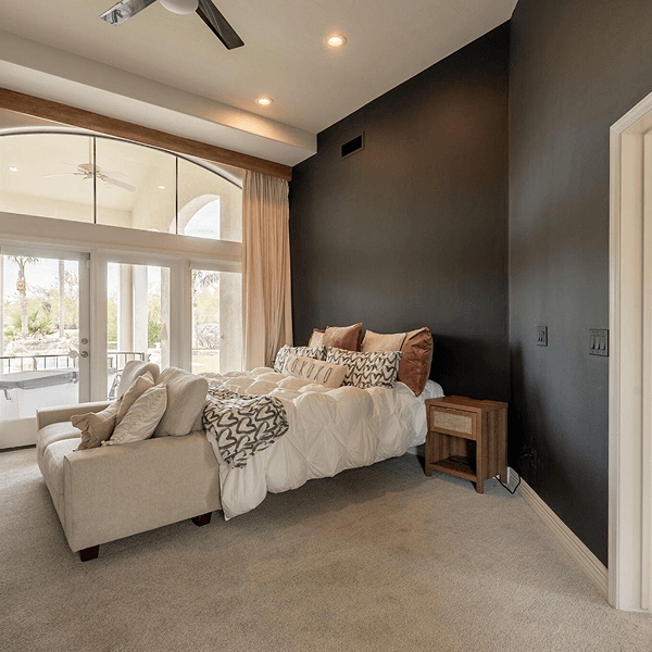 A bright, modern bedroom featuring a large bed with pillows, a chaise lounge, a dark gray accent wall, and an expansive window with outdoor views.