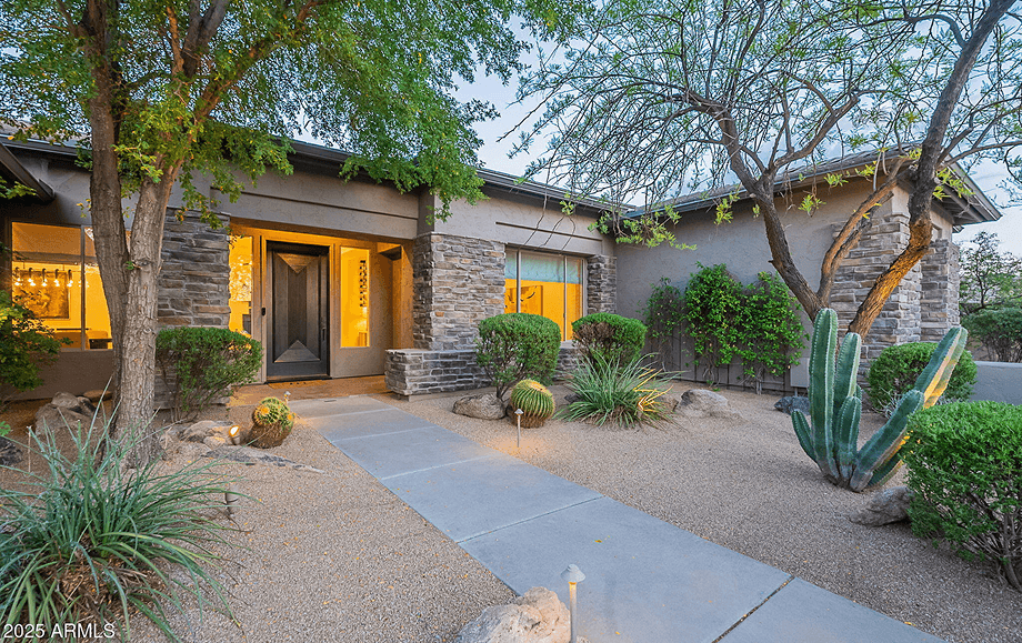 Inviting front entrance of a modern desert home at dusk, featuring a stucco and stone facade, warm interior lights, a concrete path, and drought-tolerant landscaping with cacti and trees.