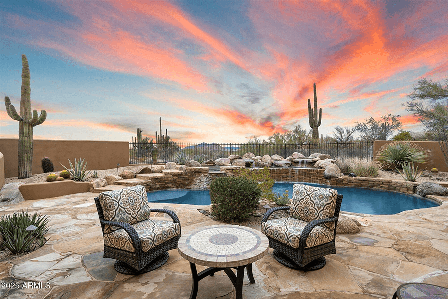 Desert backyard patio with a pool, waterfall, two chairs, and a table, surrounded by saguaro cacti and desert plants, under a vibrant orange and blue sunset sky.