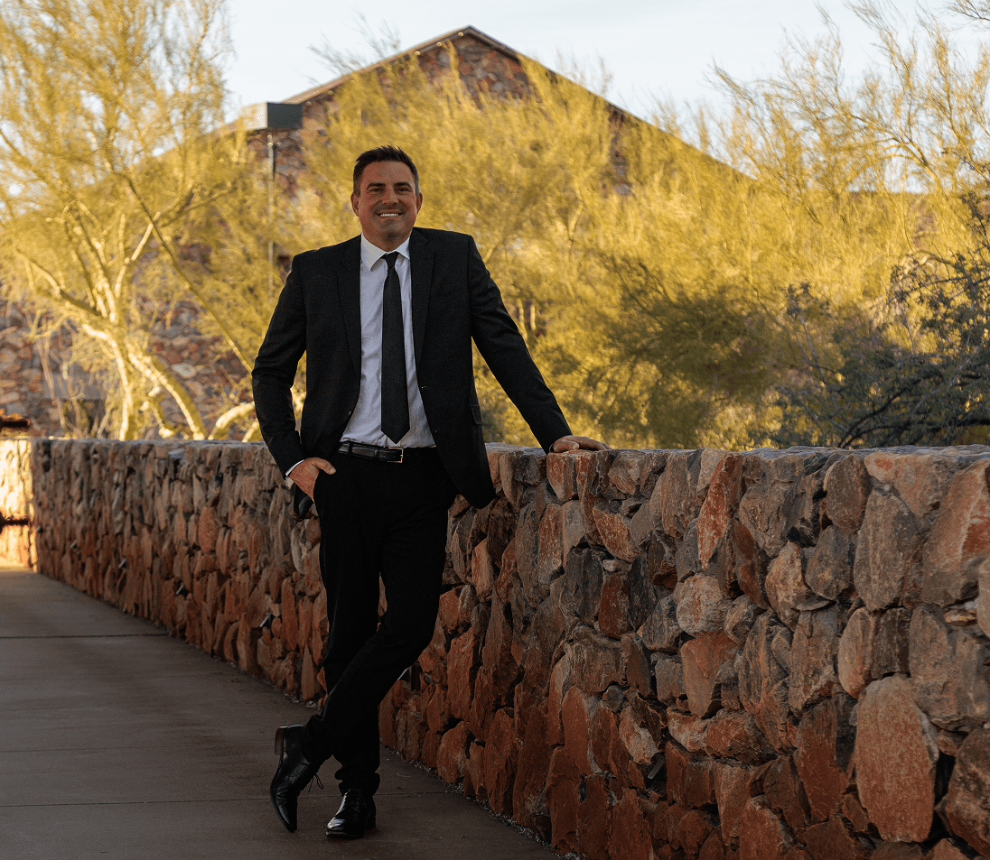 A professional man in a dark suit and tie walks through a sunlit modern hallway with large windows, looking out to the right.