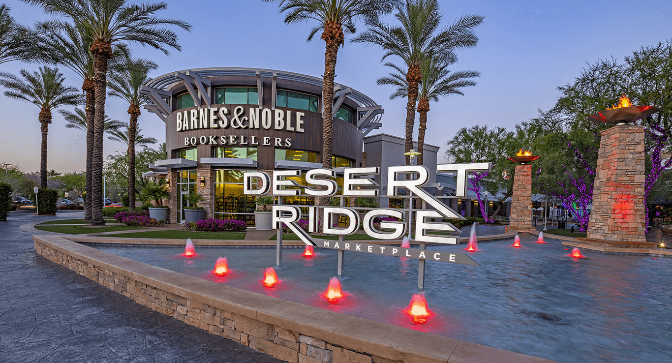 Outdoor shopping and dining area at dusk, featuring restaurants like Islands and Zupas, vibrant neon signs, palm trees, and people strolling through the illuminated promenade.
