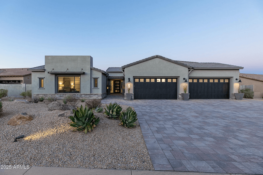 Modern single-story home at dusk with a light gray exterior, dark garage doors, desert landscaping, and a paver driveway, illuminated by interior and exterior lights.