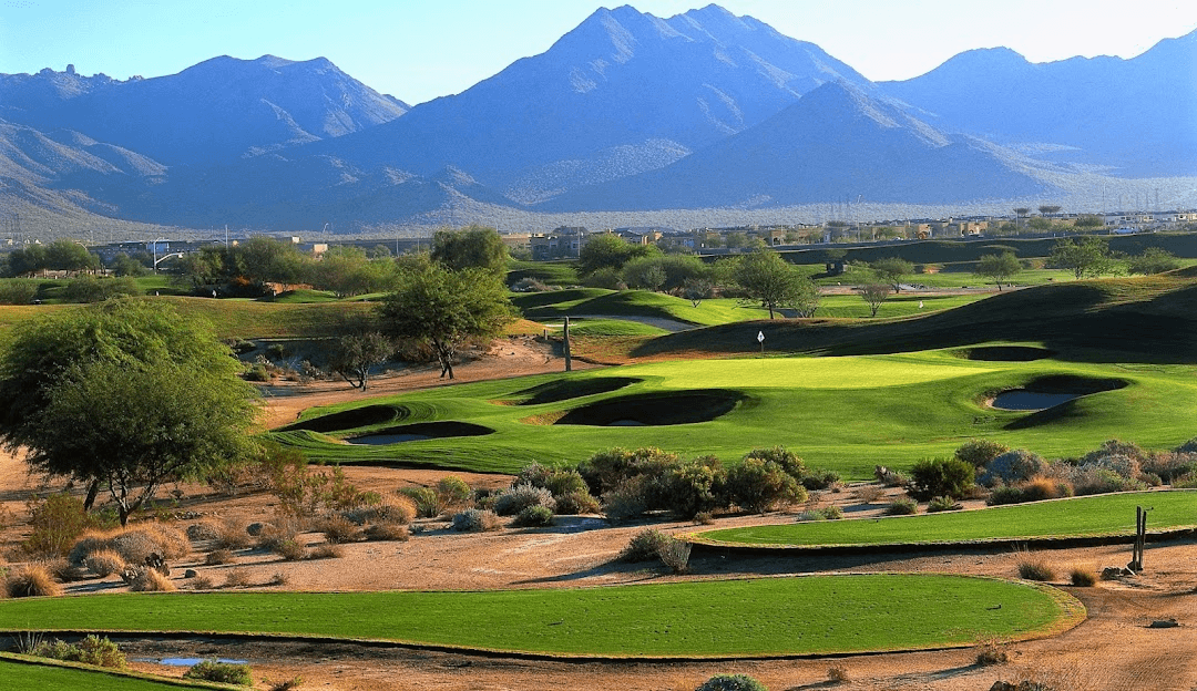 A vibrant golf course with green fairways, bunkers, and a flagstick, set against a backdrop of imposing blue mountains under a clear sky, with desert vegetation in the foreground.