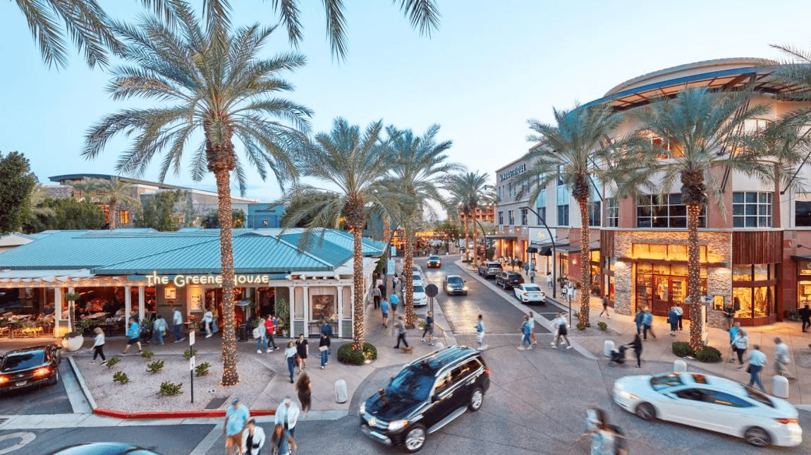 Lively outdoor shopping district at dusk, featuring palm trees, bustling pedestrians, cars, and visible storefronts including 'The Greene House'.