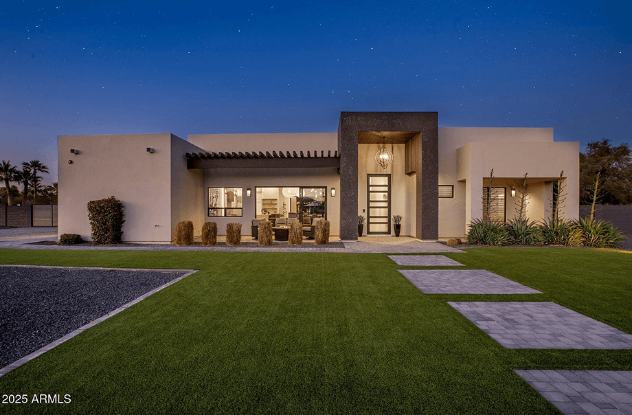 Modern single-story house exterior at night with illuminated interior, light stucco walls, and a dark stone entryway. A manicured lawn and paver pathway are in the foreground under a starry sky.