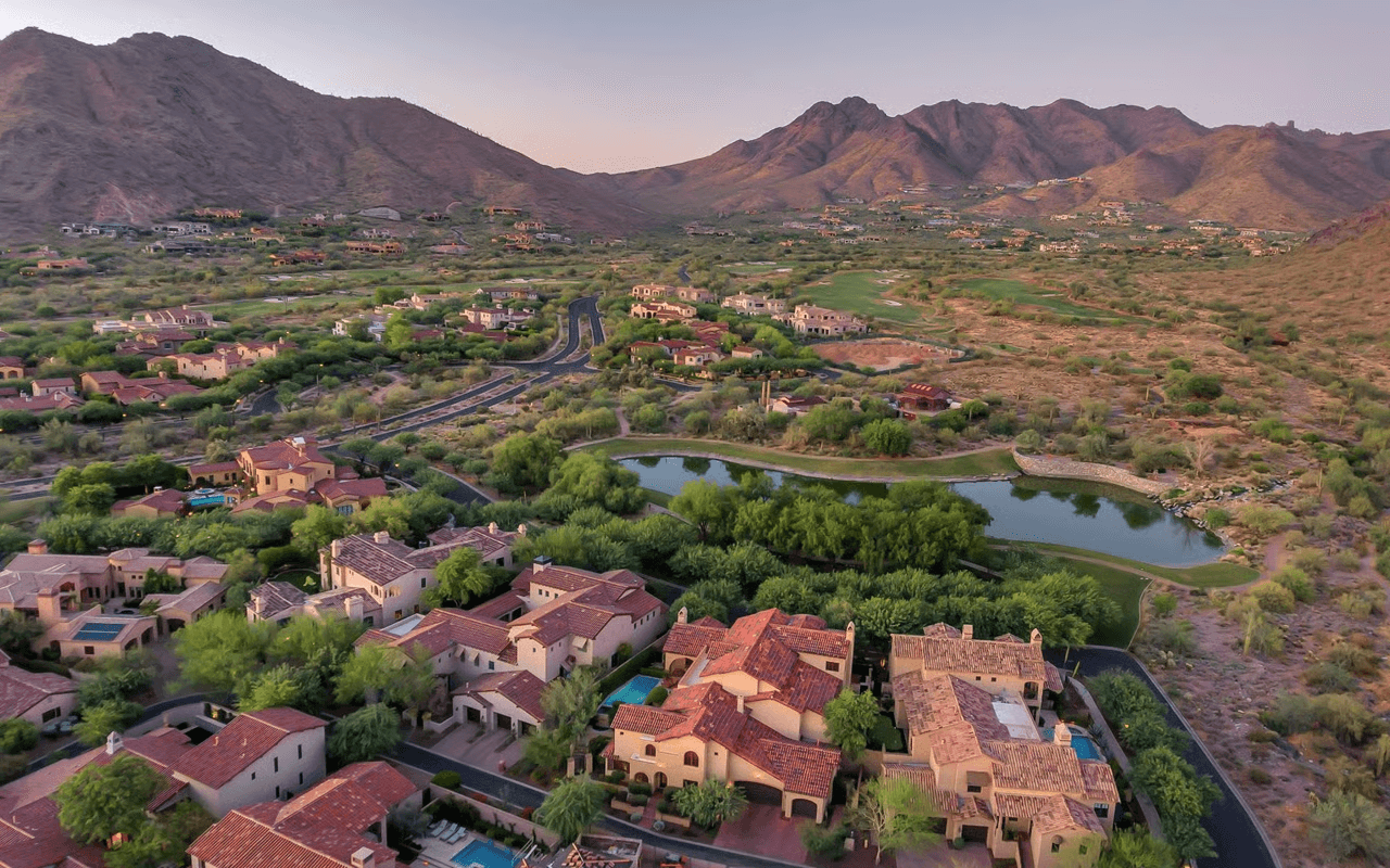 Aerial view of a luxury residential community with red-roofed houses, a golf course, and a pond, nestled in a rugged desert mountain landscape at dusk.