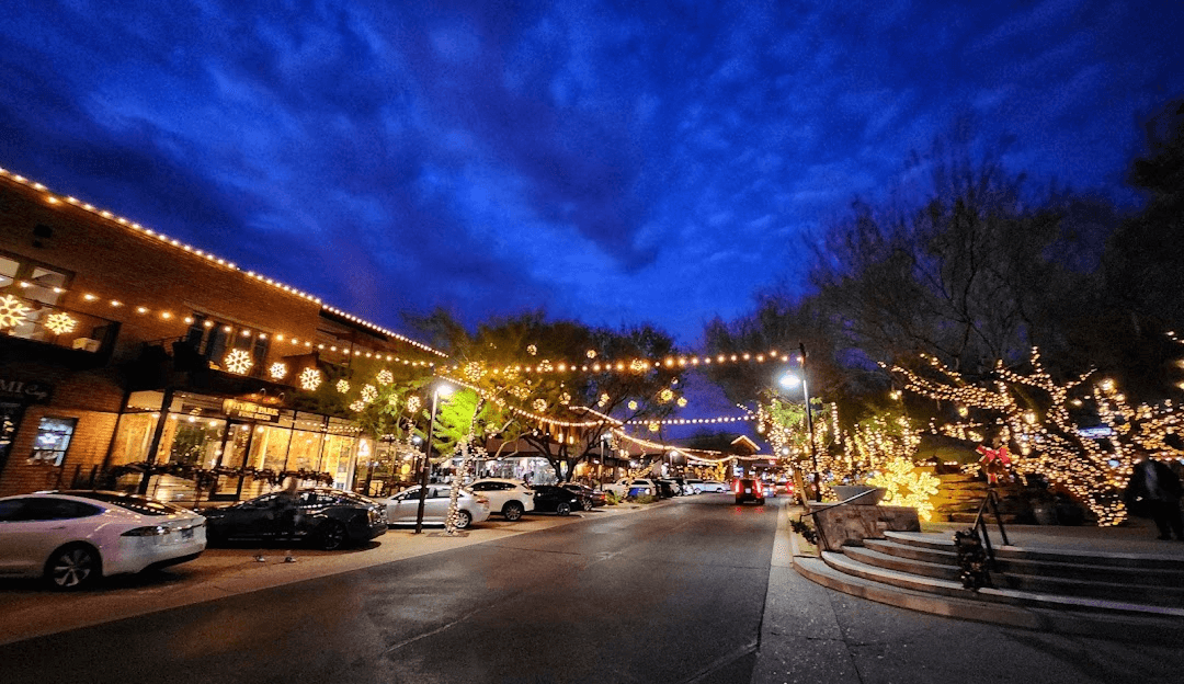 A festive street scene at dusk, with buildings and trees adorned with numerous warm string lights and large snowflake decorations, creating a cheerful atmosphere.