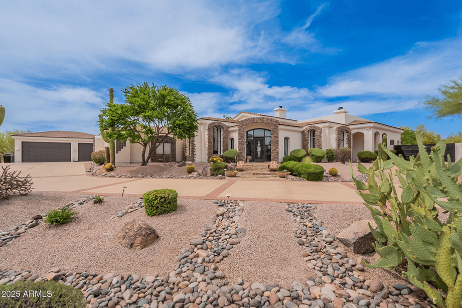 Luxury desert home with cream stucco walls, stone arches, a large entry, and a circular driveway surrounded by desert landscaping and cacti under a blue sky.