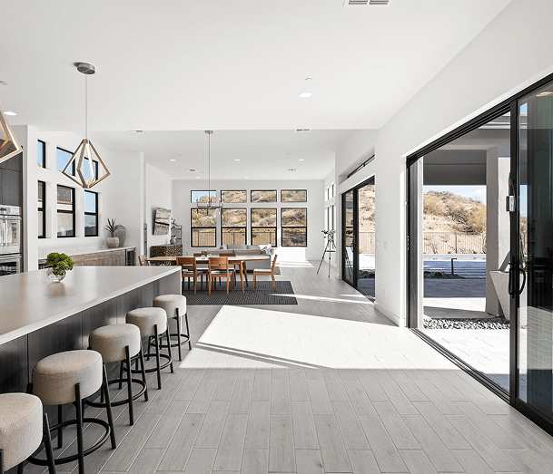 A modern open-concept home interior with a kitchen island, dining area, and living room, featuring large windows and sliding doors opening to a desert landscape.