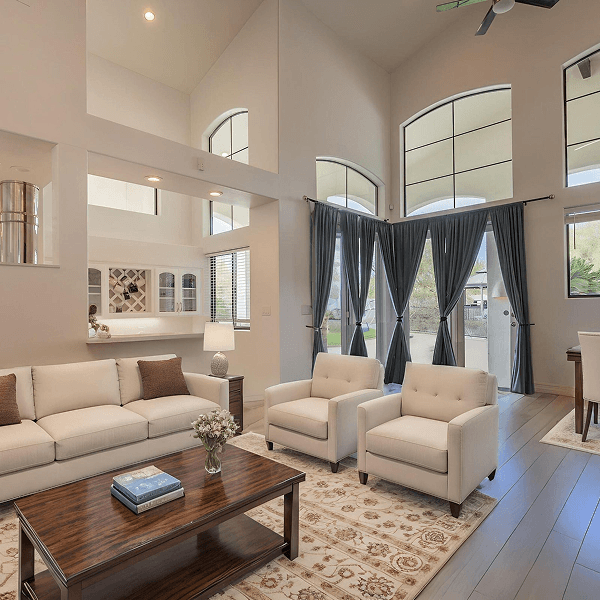 A spacious modern living room with a white sofa, two cream armchairs, and a dark wood coffee table on a patterned rug. Large arched windows with grey curtains overlook an outdoor patio, and a built-in bar area is in the background.