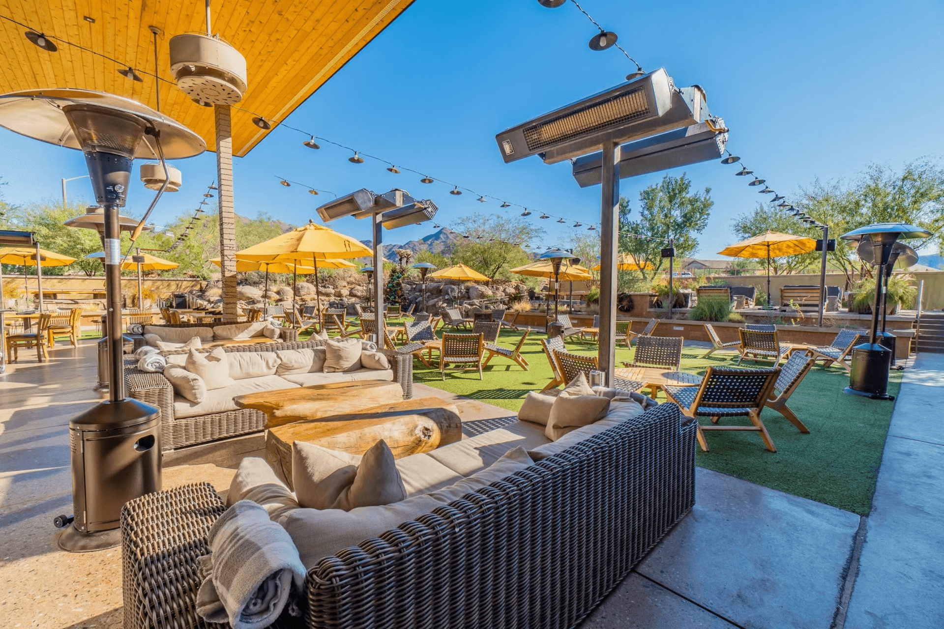 An expansive outdoor patio lounge area featuring wicker sofas, wooden tables, yellow umbrellas, string lights, and patio heaters under a clear blue sky, with a desert landscape background.