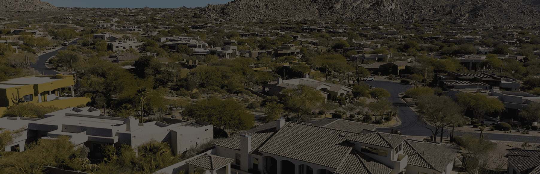 Aerial view of an upscale residential community set within a desert landscape, featuring numerous houses, winding roads, and desert flora, with rocky mountains in the background.