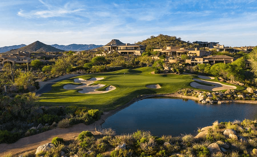 A scenic desert golf course green with sand traps and a lake, surrounded by luxury homes nestled into hills and mountains under a blue sky.